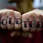 Фотография татуировки на костяшках пальцев: надпись 'HOLD THEM' выполненная в традиционном стиле с использованием красного и черного цветов.