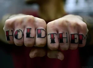 Фотография татуировки на костяшках пальцев: надпись 'HOLD THEM' выполненная в традиционном стиле с использованием красного и черного цветов.
