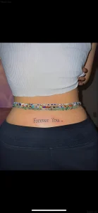 Фотография татуировки на пояснице с надписью 'Forever You' выполненной тонким шрифтом, расположенной горизонтально между бедрами.