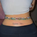 Фотография татуировки на пояснице с надписью 'Forever You' выполненной тонким шрифтом, расположенной горизонтально между бедрами.