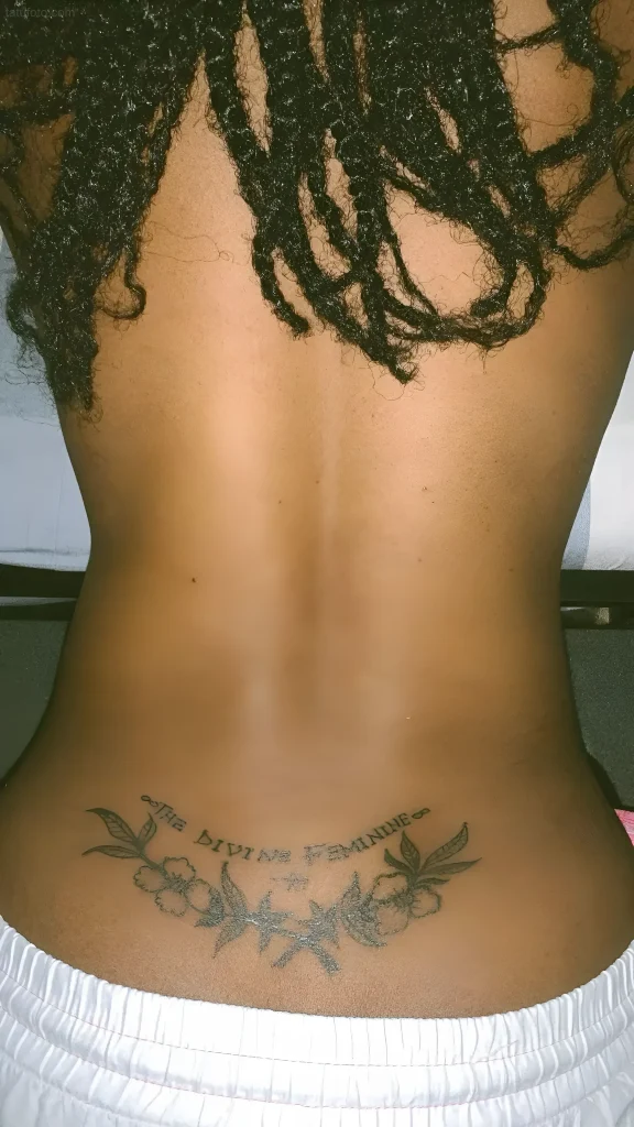 Photo of a woman's lower back tattoo: delicate flowers and leaves framing the inscription 'The Divine Feminine' in minimalist style.