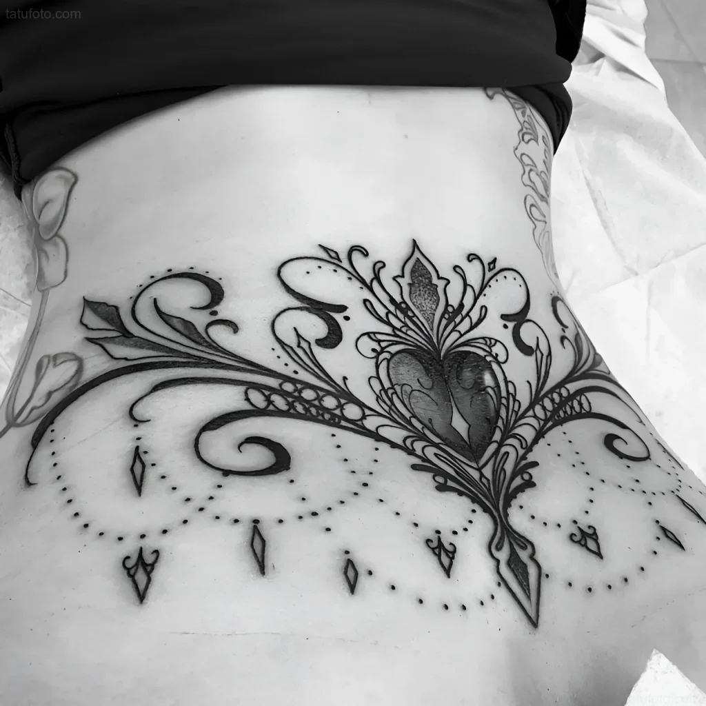 Black and white lower back tattoo with a heart surrounded by decorative floral elements and dots, done in a vintage style.