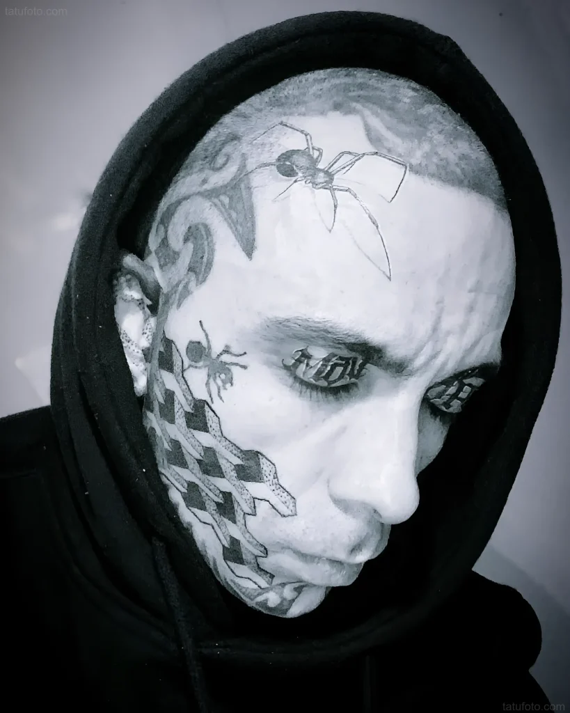 Black and white photo of a man with spider tattoos on his eyelids and geometric patterns on his face, showcasing an extreme tattooing style.