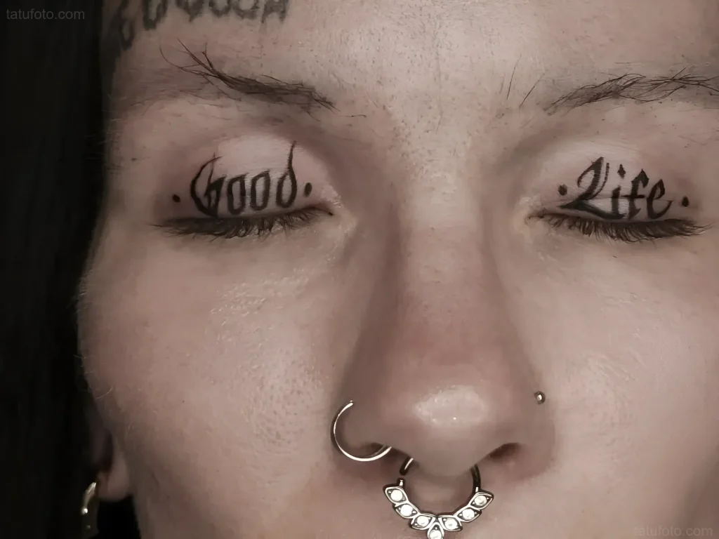 Black and white eyelid tattoo with 'Good' and 'Life' inscriptions under the lashes, done in a minimalist style.