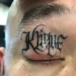 Черно-белая татуировка имени "Kipke" готическим шрифтом над глазом мужчины, выполненная на веке.