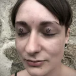Фотография девушки с двумя небольшими геометрическими татуировками на веках, выполненными в технике микротату, и тремя точками на лбу.