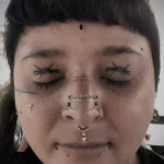 Фотография девушки с татуировкой на веках, имитирующей закрытые глаза, и множеством пирсинга на лице.
