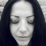 Черно-белое фото девушки с татуировкой в виде нескольких маленьких точек под каждым глазом, подчеркивающих выразительность взгляда.
