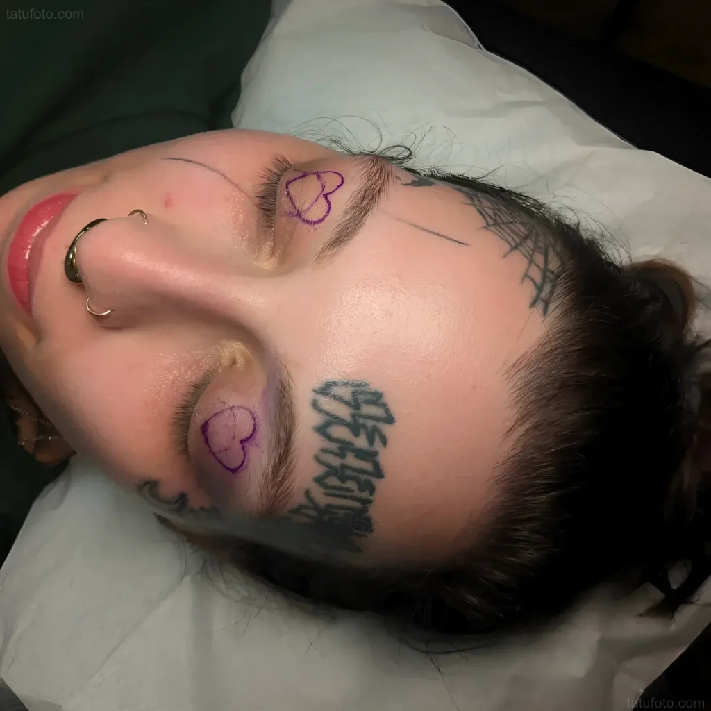 Photo of an eyelid tattoo: inscription and small hearts, done in black, on a girl's face with piercings.