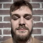 Мужчина с татуировками на веках, надписи 'FEAR GOD' над глазами, борода, пирсинг носа, кирпичная стена на фоне.
