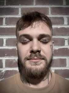 Мужчина с татуировками на веках, надписи 'FEAR GOD' над глазами, борода, пирсинг носа, кирпичная стена на фоне.