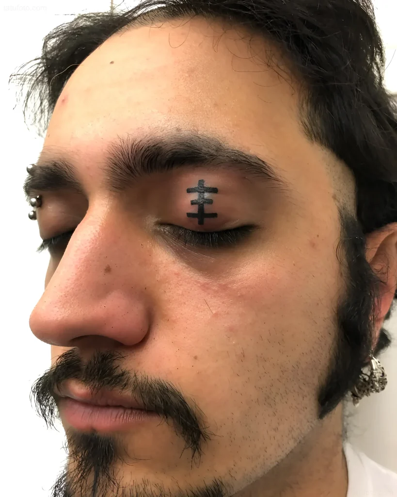眼睑纹身照片：一个留着胡须、戴着穿孔的男人的脸上有一个小黑色十字架，采用图形风格。