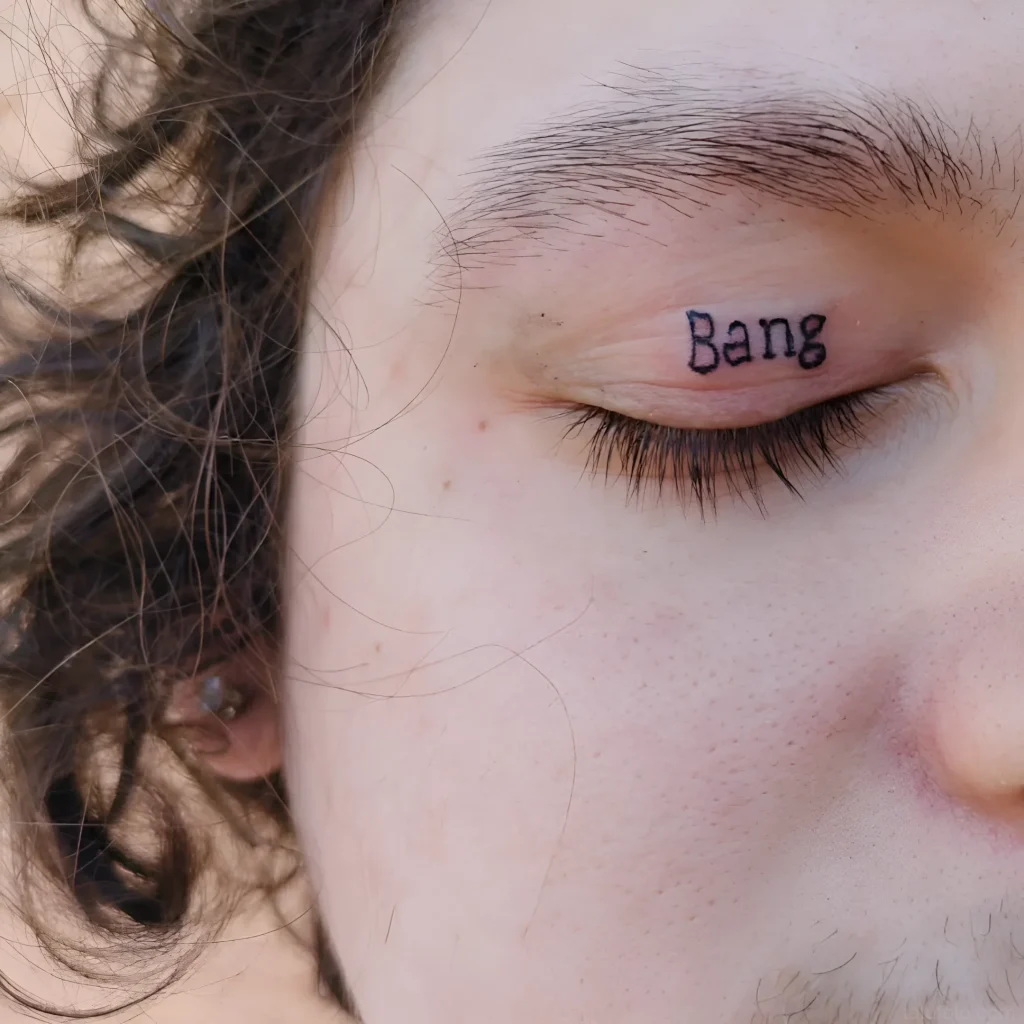 眼睑纹身特写，上面写着“Bang”字样，黑色，背景是浅色皮肤和卷发。