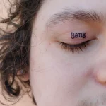 Крупный план татуировки на веке с надписью 'Bang' черным цветом, на фоне светлой кожи и кудрявых волос.