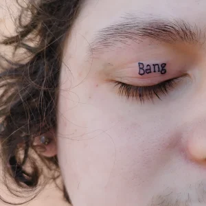 Крупный план татуировки на веке с надписью 'Bang' черным цветом, на фоне светлой кожи и кудрявых волос.
