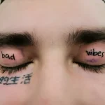 Фото татуировки на веках: надписи 'bad vibes' и иероглифы под глазами, выполненные черными чернилами, на мужском лице.