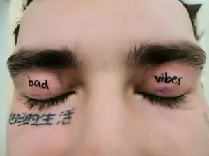 Фото татуировки на веках: надписи 'bad vibes' и иероглифы под глазами, выполненные черными чернилами, на мужском лице.