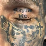 Крупный план татуировки на веке с надписью 'Forgive Me' и фрагментом татуировки креста на щеке.
