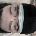 Черно-белая татуировка на веках в виде тонких колючих лиан, расположенных над бровями, на фоне светлой кожи и медицинской маски.