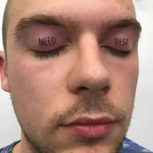 Фотография татуировки на веках мужчины: надписи 'NEED' на левом веке и 'REST' на правом, выполненные черным цветом в минималистичном стиле.
