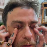 Фотография мужчины с татуировкой иероглифа на верхнем веке, выполненной в технике тонкой линии. Дополнительные пирсинги на лице и татуировки на руках.