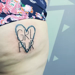 Фотография татуировки на ягодице: черная надпись 'GRL PWR' заключена в контур сердца, выполненная в минималистичном стиле.