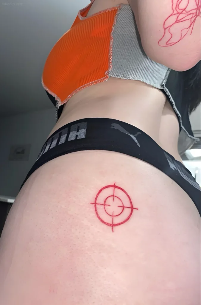 Photograph of a tattoo of a red crosshair on a girl's buttock, in a minimalist style.