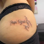 Фото татуировки на ягодице: черная надпись 'Papi Sigue' и минималистичный рисунок женского профиля, выполненные в технике тонкой линии.