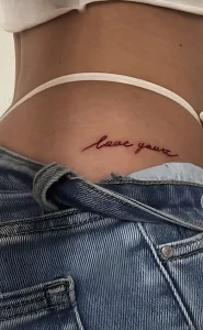 Фото татуировки на ягодицах: надпись 'love yourz' красными чернилами, выполненная каллиграфическим шрифтом на нижней части спины девушки.