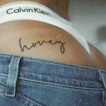 Минималистичная татуировка надпись 'honey' черными чернилами на ягодице девушки в джинсах и белом белье