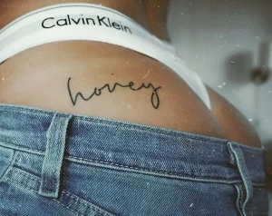 Минималистичная татуировка надпись 'honey' черными чернилами на ягодице девушки в джинсах и белом белье