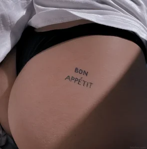 Фото татуировки на ягодице: надпись 'Bon Appétit' выполненная тонкой линией, минималистичный стиль, женственное тело.