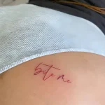 Фотография татуировки надписи 'Bite Me' на ягодице, выполненной в технике микрореализма, с акцентом на изящный шрифт и нежный розовый оттенок.