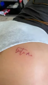 Фотография татуировки надписи 'Bite Me' на ягодице, выполненной в технике микрореализма, с акцентом на изящный шрифт и нежный розовый оттенок.