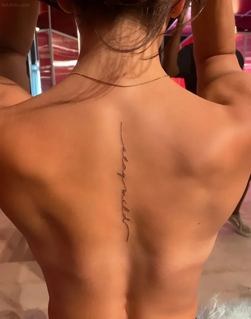 Photograph of a girl's back tattoo: the inscription 'be wild' along the spine, done in a fine line.