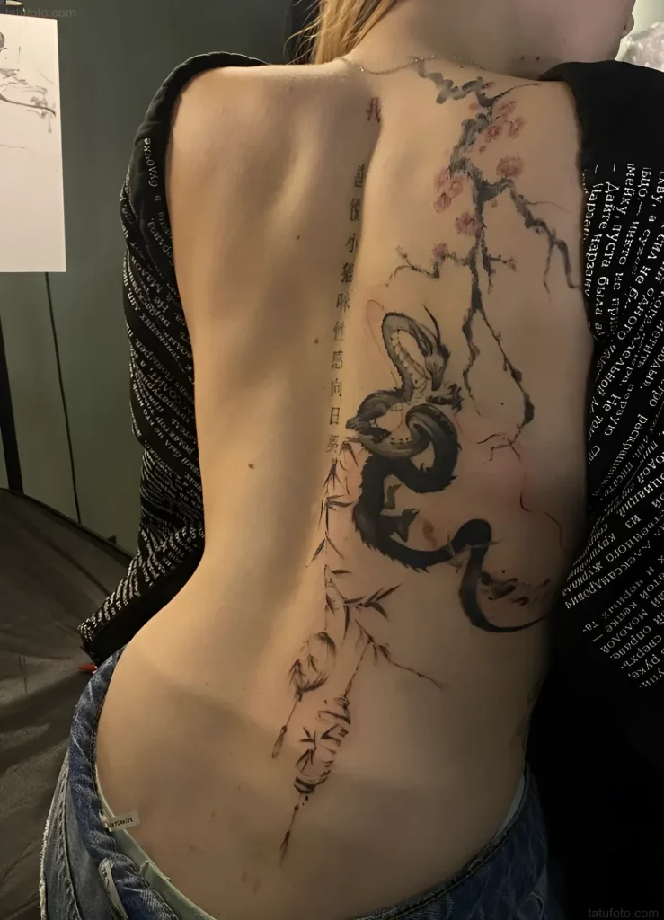 Tattoo photo along the spine: a black dragon coils around a sakura branch against a background of ancient text, side view.