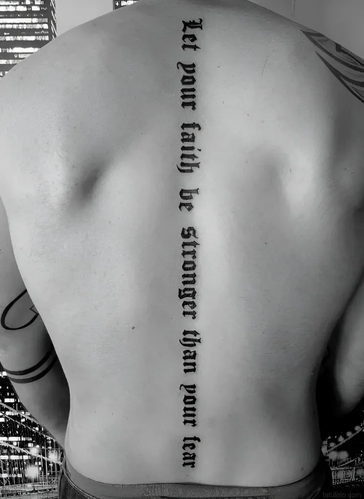 Black and white photo of a tattoo along a man's spine with the inscription in Gothic font 'Let your faith be stronger than your fear'.