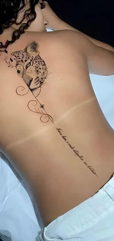 Photo of a tattoo: a graphic-style portrait of a leopard's head, done along a girl's spine, with added decorative elements and a Latin inscription.