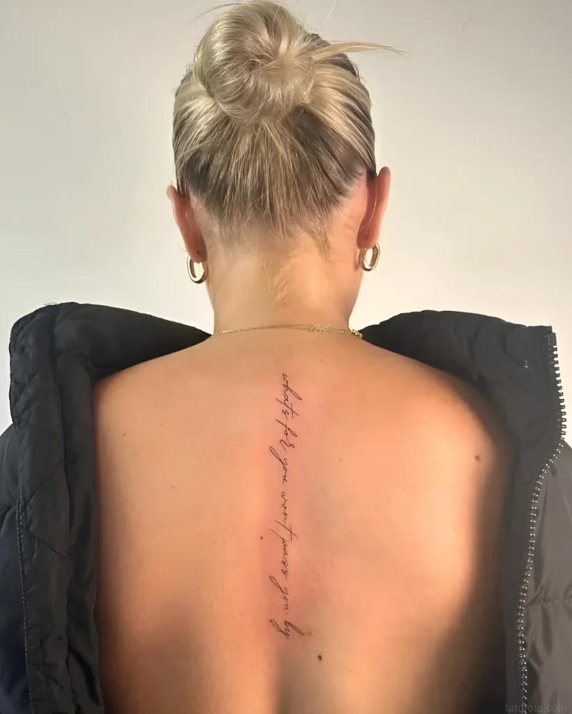 Photograph of a girl's back tattoo - a thin inscription in a calligraphic font, located along the spine, symbolizing a personal story and meaningful words.