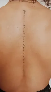 Фотография татуировки вдоль позвоночника с надписью 'Dream without fear, Love without limits' выполненной тонкой линией, на загорелой женской спине.