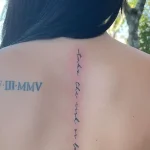 Фото татуировки на спине девушки: вертикальная надпись 'Take the risk or lose the chance' вдоль позвоночника и римские цифры VII-III-MMV на лопатке.