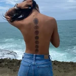 Фотография девушки с татуировкой семи чакр, расположенных вдоль позвоночника, на фоне моря. Детализированное изображение символов чакр.