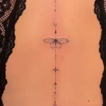 Фотография татуировки на спине женщины: тонкая линия тату с изображением бабочки и геометрических символов, расположенная вдоль позвоночника.