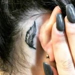 Фотография татуировки: небольшое перо, выполненное в технике blackwork, расположено за ухом у девушки с темными волосами.