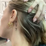 Фотография татуировки за ухом: три черные цифры '7' расположены вертикально на шее девушки, минималистичный дизайн.