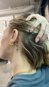 Фотография татуировки за ухом: три черные цифры '7' расположены вертикально на шее девушки, минималистичный дизайн.