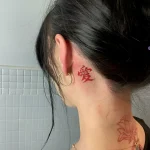 Крупный план татуировки за ухом: красный иероглиф 'Любовь' на нежной женской коже, минималистичный стиль.
