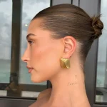 Фотография татуировки за ухом: изящная надпись 'lover' курсивом и маленький крестик на шее девушки с собранными волосами.