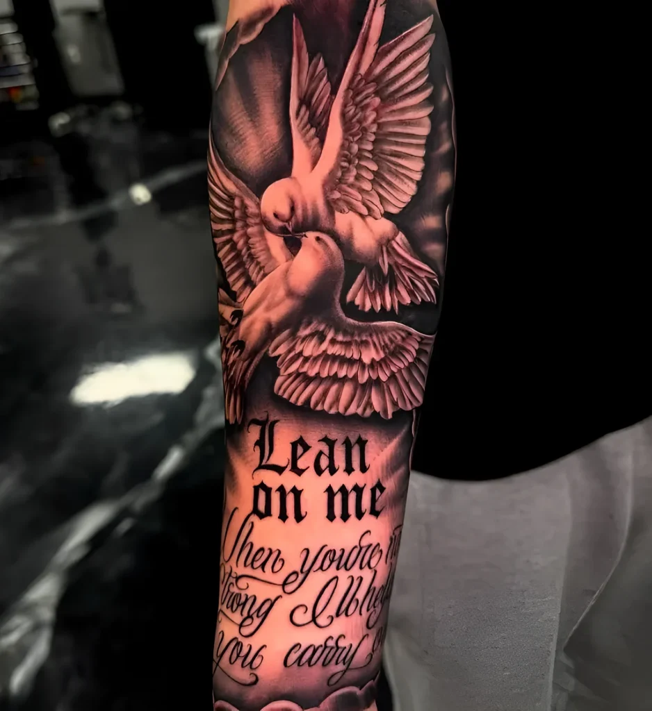 Black and white forearm tattoo depicting two doves taking flight into the sky, with the calligraphic inscription 'Lean on me'.