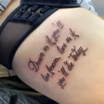 Фотография татуировки на боку женщины: изящная надпись "Dream as if you'll live forever, live as if you'll die today" выполненная тонкой линией.